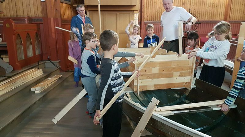 To voksne og mange barn står rundt en robåt (Noahs Ark) og bygger et hus på båten