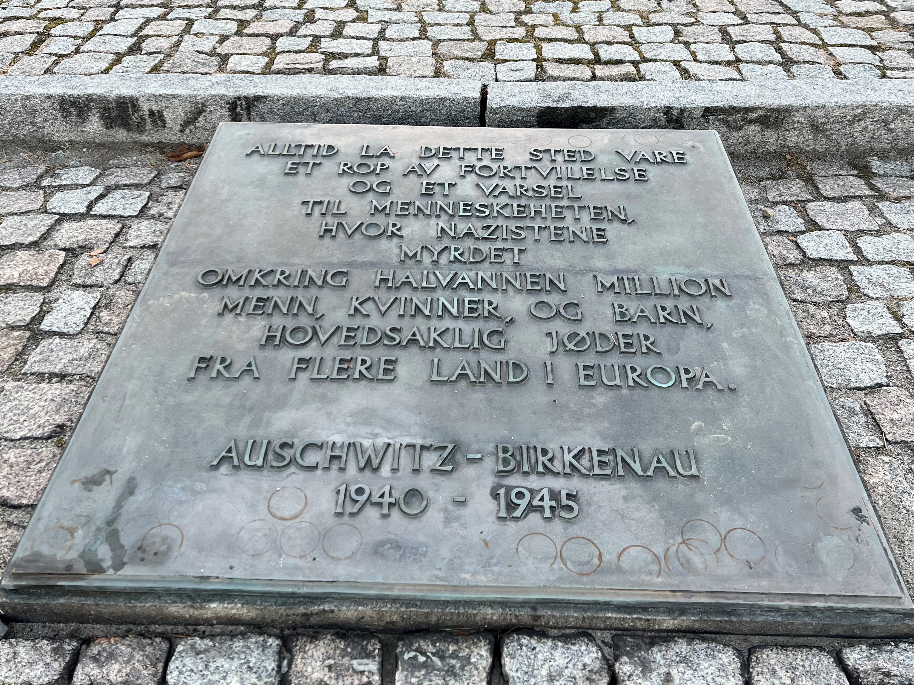 Plakett med tekst: Alltid la dette sted være et rop av fortvilelse og et varsel til menneskeheten, hvor nazistene myrdet omkring halvannen million menn, kvinner, og barn, hovedsaklig jøder fra flere land i Europa. Auschwitz-Birkenau 1940-1945.