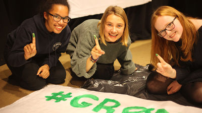 Tre jenter maler på et banner med grønnfarge. De viser frem pekefingene med maling på.